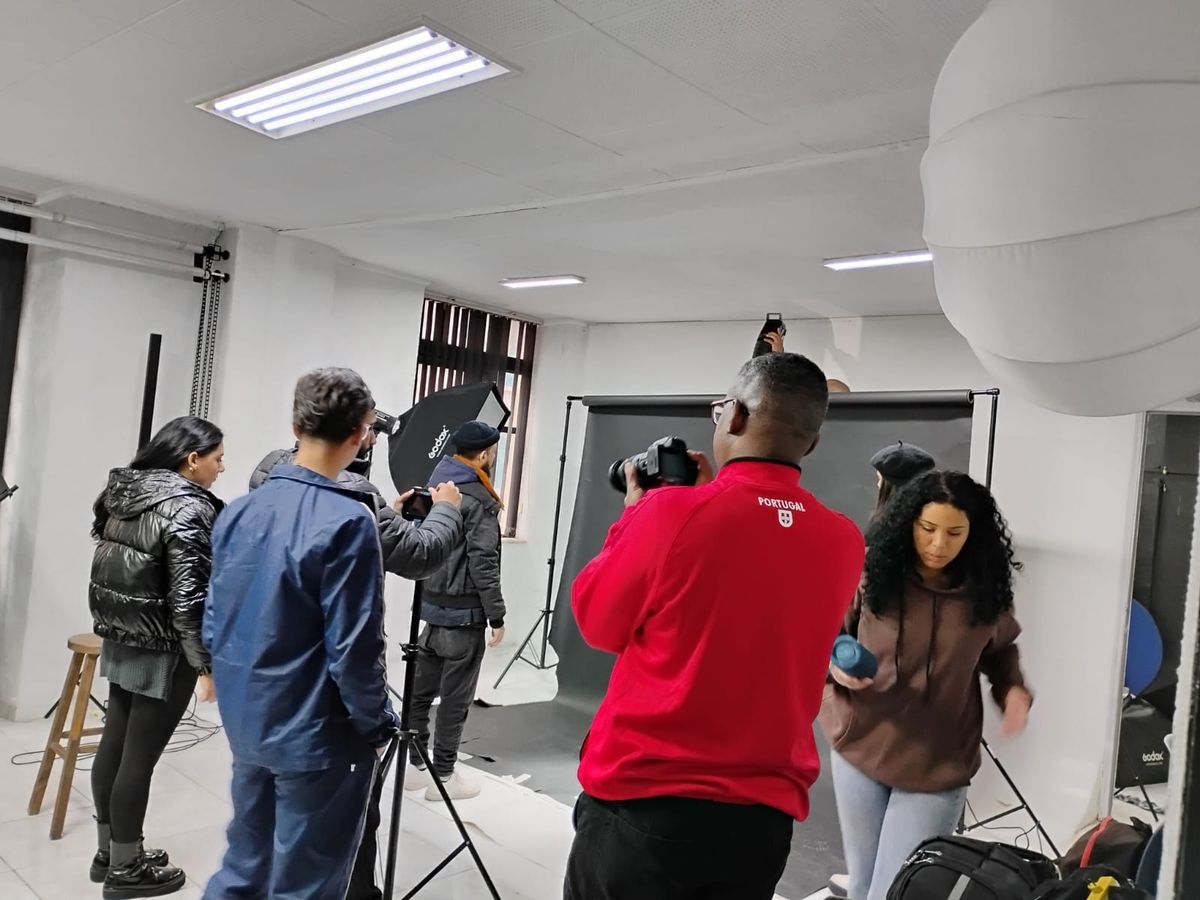 Bastidores making Off. Imagem de um estúdio fotográfico com paredes e teto branco, da esquerda para direita temos: uma mulher vestida de preto, a frente da mulher um homem vestido de preto mexendo na câmera, a frente do homem do lado esquerdo outro homem vestido de preto e laranja mexendo na luz, ao lado direito outros dois homens um vestido de azul e um homem vestido de calças preta e blusa vermelha de frio ao seu lado direito uma mulher anda em direção a câmera, vestida de blusa de frio marrom e calças jeans, a estação do ano é o inverno.