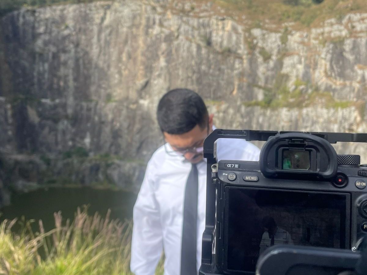 Bastidores making Off. Imagem de um homem vestido de branco, com óculos e uma gravata preta ao fundo de uma pedreira, na frente do homem à direita uma câmera fotográfica saindo de um enquadramento.