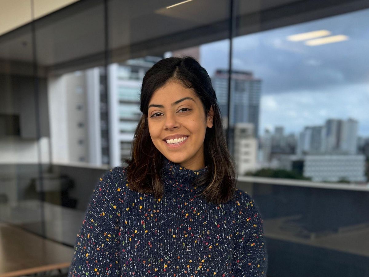Na foto vemos Danielle Carvalho, mulher morena com cabelos castanhos lisos na altura dos ombros, ela está com uma blusa de frio azul e sorri, ao fundo uma imagem de prédios fora de foco.